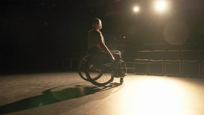 A person seated in a wheelchair is spotlighted on a stage.