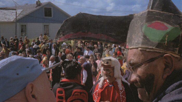 In the foreground of a crowd, a man wearing a ceremonial mask-helmet and glasses appears to be in discussion with another man wearing a blue cap.